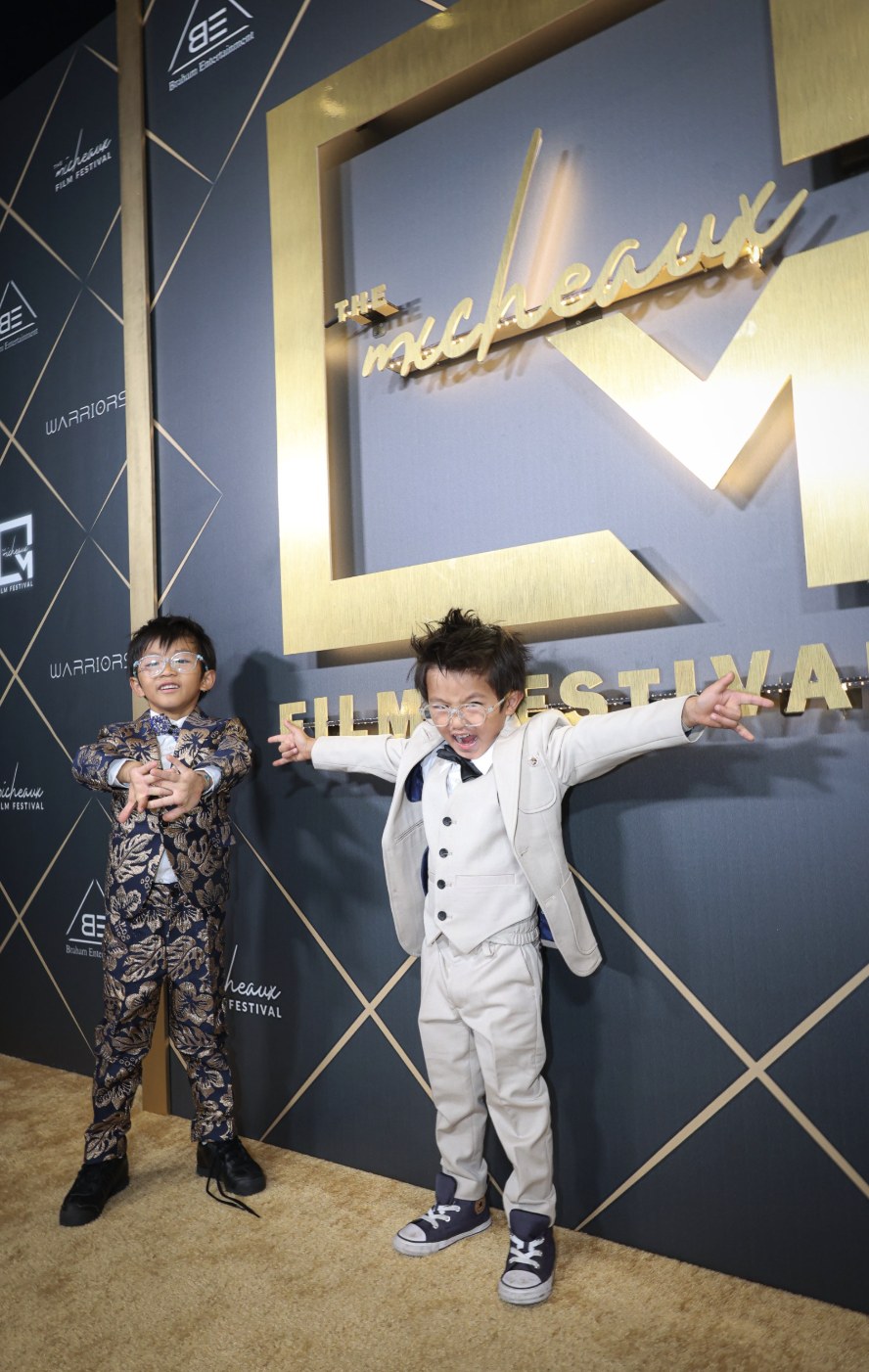 Two young guests in suits striking triumphant poses on the gold Micheaux Film Festival carpet.