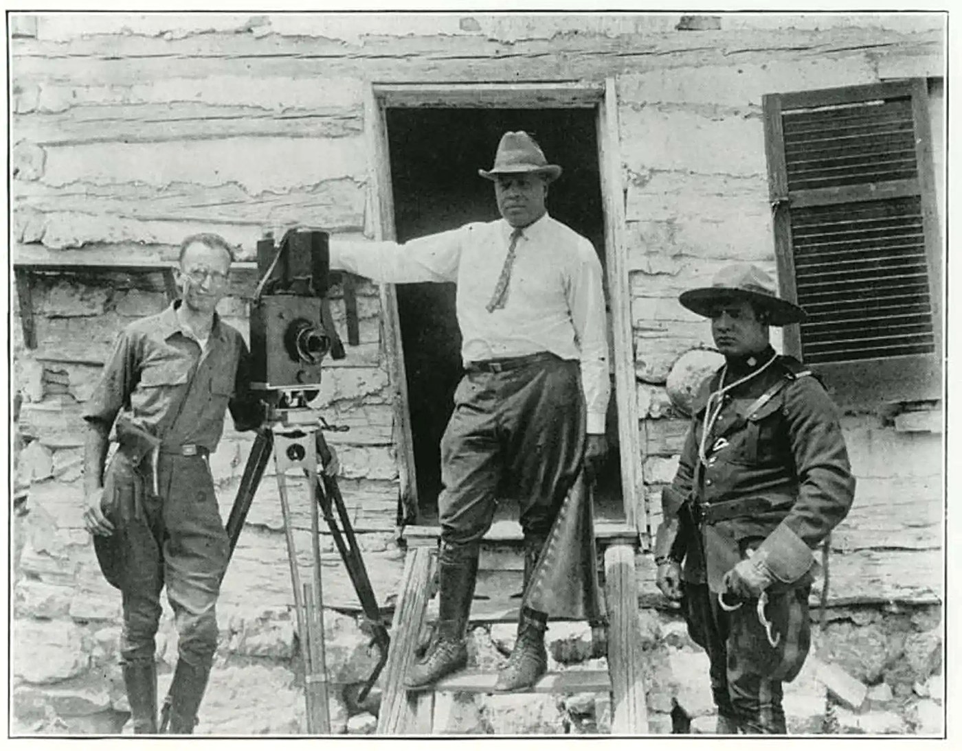 Oscar Micheaux on location, standing in a doorway with his hand resting on a film camera, flanked by his crew. Archival photograph, circa 1920s.